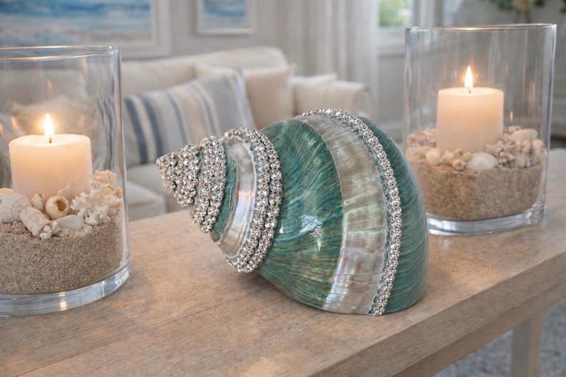 May include: A decorative seashell with a teal and white iridescent finish, embellished with rows of clear rhinestones. It is positioned between two clear glass cylindrical candle holders, each containing a white pillar candle, sand, and shells.