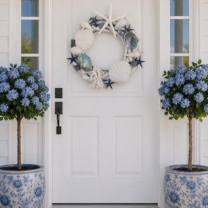 May include: A white front door adorned with a coastal wreath featuring seashells, starfish, and coral. Flanking the door are two blue and white floral planters with small trees, creating a welcoming entrance.
