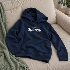 Könnte beinhalten: Ein dunkelblauer Kapuzenpullover mit langen Ärmeln und dem weißen Schriftzug "MAMAS Spätzle" auf der Brust. Das Kleidungsstück verfügt über eine Kapuze, lange Ärmel mit gerippten Bündchen sowie einen gerippten Saum aus einfarbigem Stoff.