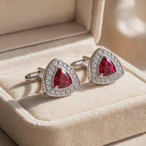 Peut inclure: Une paire de boutons de manchette argentés avec une pierre précieuse rouge de forme triangulaire au centre, entourée d'une bordure de petites pierres claires et scintillantes. Les boutons ont une finition métallique et sont présentés dans un écrin.