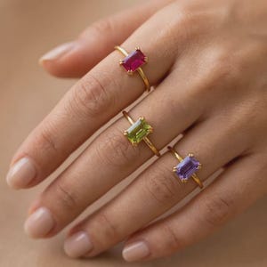 May include: Three gold rings, each featuring a single rectangular-cut gemstone. The top ring has a vibrant pink stone, the middle ring holds a light green stone, and the bottom ring displays a soft purple stone, all set on thin gold bands.