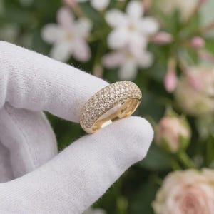 May include: A gold band ring featuring a wide, domed surface completely encrusted with numerous small, clear, sparkling stones arranged in a dense, uniform pavé setting, showcasing a high-shine metallic finish and a rounded, substantial profile.