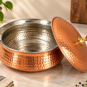 May include: A round serving bowl with a hammered copper exterior and a smooth, polished stainless steel interior. The bowl includes a matching hammered copper lid with a shiny, gold-toned brass knob handle centered on top for easy lifting.