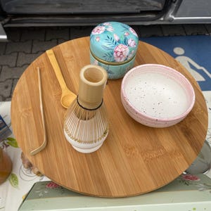 Könnte beinhalten: Ein rundes Bambustablett mit einem Bambus-Matchabesen, einem Bambuslöffel, einer Bambus-Teeschaufel, einer hellrosa gesprenkelten Keramikschale und einer kleinen türkisfarbenen Dose mit rosa Blumenmuster auf einer flachen, runden Holzoberfläche.