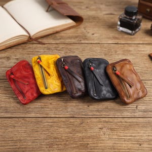 Puede incluir: Cinco pequeñas carteras de cuero en rojo, amarillo, marrón oscuro, negro y tostado. Cada cartera rectangular cuenta con un bolsillo vertical con cremallera y una lengüeta de cuero con una banda roja. La cartera tostada tiene las letras A y B.