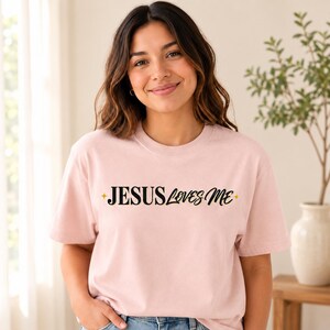 Puede incluir: Una camiseta de manga corta de color rosa claro con el texto negro "JESUS LOVES ME" impreso en el pecho. El diseño incluye dos pequeños iconos de estrellas doradas de cuatro puntas, uno antes de la palabra "JESUS" y otro después de "ME".