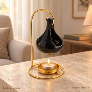 May include: A gold-toned metal stand with a circular base and a curved arm that suspends a glossy black ceramic oil warmer. A small, round metal tray sits at the base, holding a lit white tea light candle directly beneath the black ceramic vessel.
