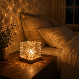 Puede incluir: Una lámpara de mesa cuadrada de cristal con una superficie texturizada y cristalina que emite una luz cálida y dorada. La lámpara está montada sobre una base de madera clara y plana, proyectando patrones de luz complejos y acuáticos.