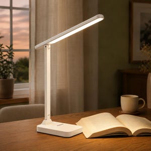 Pode incluir: Uma luminária de mesa LED branca com base retangular e braço fino ajustável. A luminária possui um painel de luz plano e alongado que está aceso, projetando um brilho intenso sobre a superfície de madeira com um livro aberto e uma caneca.
