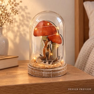 Op de afbeelding: Een glazen stolp op een ronde houten voet met een illustratie van een vos en twee grote oranje paddenstoelen. Het geheel is gevuld met lichtgekleurde houtkrullen en verlicht door dunne, warmgekleurde lichtsnoeren die om de figuren zijn gewikkeld.