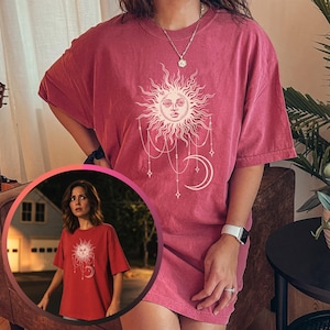 May include: A red short-sleeved t-shirt featuring a white graphic print of a sun with a face and a crescent moon connected by hanging chains and stars. The shirt has a crew neckline and a relaxed fit, shown on a person wearing a necklace.
