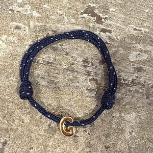 Peut inclure: Un bracelet en corde bleu marine avec des accents de points blancs. Le bracelet est orné d'un charm en forme de lettre "G" de couleur or. Le bracelet est présenté sur une surface texturée de tons neutres.
