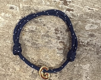 Puede incluir: Una pulsera de cuerda azul marino con detalles de puntos blancos. La pulsera presenta un dije dorado con la letra "G". La pulsera se muestra sobre una superficie texturizada de tonos neutros.