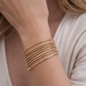 Könnte beinhalten: Ein goldfarbenes, mehrsträngiges Armband, das aus einer Kombination von glatten, polierten Drahtreifen und texturierten, gedrehten Kettengliedern besteht. Das Armband windet sich spiralförmig um das Handgelenk und endet in einer kleinen Kugel.