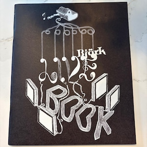 May include: A black book cover featuring white line art of a marionette controller connected by strings to the text Björk LIVE BOOK and several empty rectangular frames. The design uses intricate hatching and swirling line work throughout.