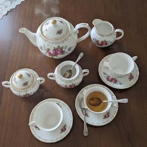 Könnte beinhalten: Ein Porzellan-Teeservice mit weißem Grund, verziert mit rosa Rosen- und blauen Blumenmustern sowie Goldrändern. Das Set besteht aus einer Teekanne, einem Milchkännchen, einer Zuckerdose mit Deckel, einer offenen Zuckerdose mit Löffel und zwei Tassen.