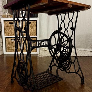 May include: A vintage Singer sewing machine base repurposed as a table, featuring a black cast iron frame with intricate scrollwork, a large side flywheel, a foot treadle, and a rectangular wooden tabletop with a dark, weathered finish.