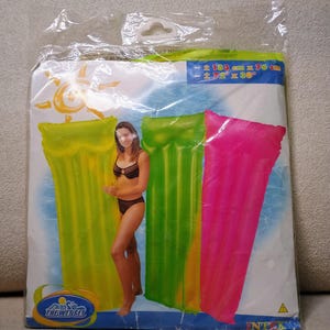 May include: A clear plastic packaging bag containing three inflatable pool floats in neon yellow, green, and pink. The packaging features a graphic of a person, a sun, and the text The Wet Set and Intex. Dimensions are listed as 72 inches by 30 inches.