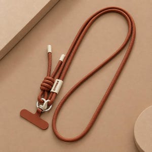 May include: A rust-colored cord lanyard featuring a long, braided rope strap, a sliding off-white plastic adjustment bead, and a matching rust-colored T-shaped connector piece attached to a silver-toned metal ring at the base of the cord.