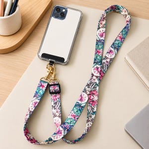May include: A smartphone in a clear protective case attached to a long fabric lanyard. The lanyard features a vibrant floral pattern with pink, teal, and white blossoms. It includes gold-toned metal swivel clasps and a black plastic slider.