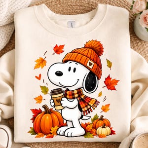 Puede incluir: Una sudadera de cuello redondo color crema con un gráfico de un perro de dibujos animados blanco que lleva un gorro de punto naranja con un corazón y una bufanda a cuadros. El perro sostiene un café, rodeado de calabazas y hojas de otoño.
