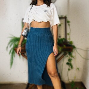 May include: A woman wearing a white cropped t-shirt with a logo, a teal knitted skirt with a side slit, and white sneakers with black stripes.