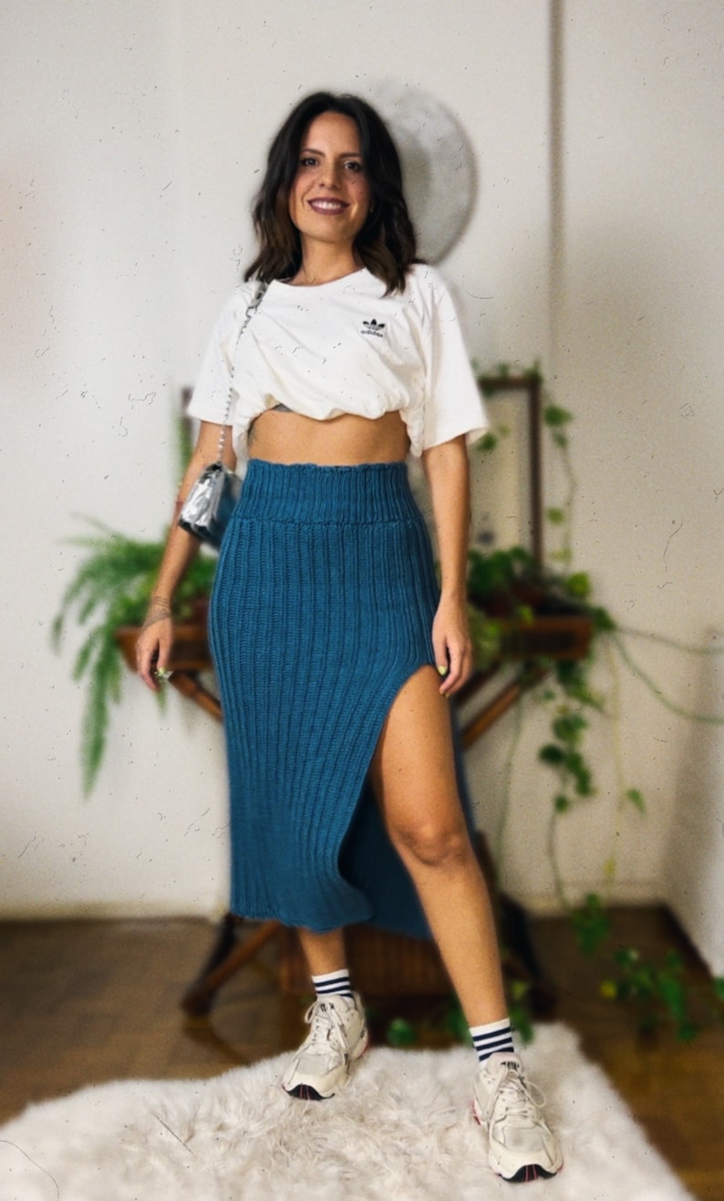 May include: A woman wearing a white cropped t-shirt with a logo, a teal knitted skirt with a side slit, and white sneakers with black stripes.