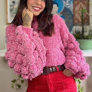 May include: A pink fluffy sweater with a high neck and large, puffy sleeves. The sweater is paired with red jeans and a brown belt.