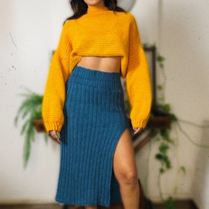 May include: A woman wearing a yellow cropped sweater and a blue knitted skirt with a slit. She is wearing black boots.