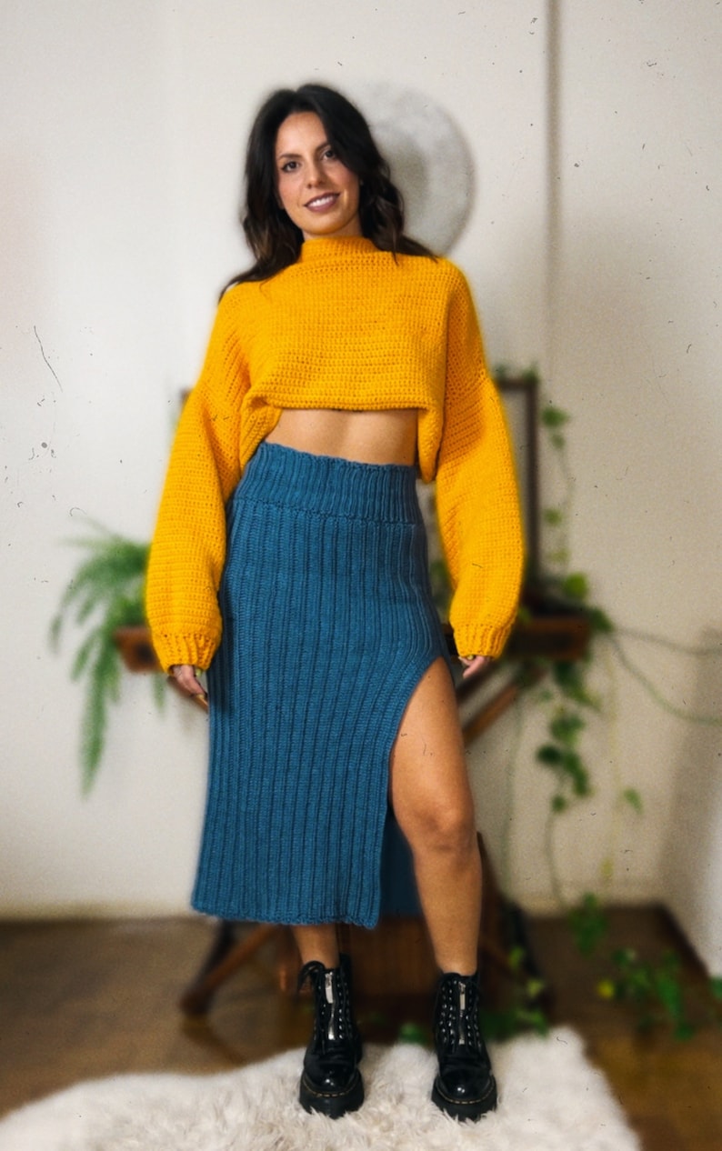 May include: A woman wearing a yellow cropped sweater and a blue knitted skirt with a slit. She is wearing black boots.