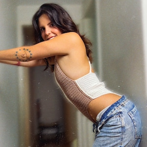 May include: A woman wearing a brown and white crocheted top and blue jeans. She has a tattoo on her arm.