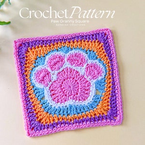 May include: A colorful crochet granny square with a pink paw print in the center. The square is made with multiple colors of yarn, including pink, orange, blue, and purple.