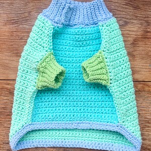 May include: A crocheted dog sweater in shades of blue, green, and light green. The sweater has a turtleneck and long sleeves.