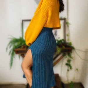 May include: A woman wearing a yellow crocheted sweater and a teal blue crocheted skirt with a slit up the side. She is wearing black leather boots.