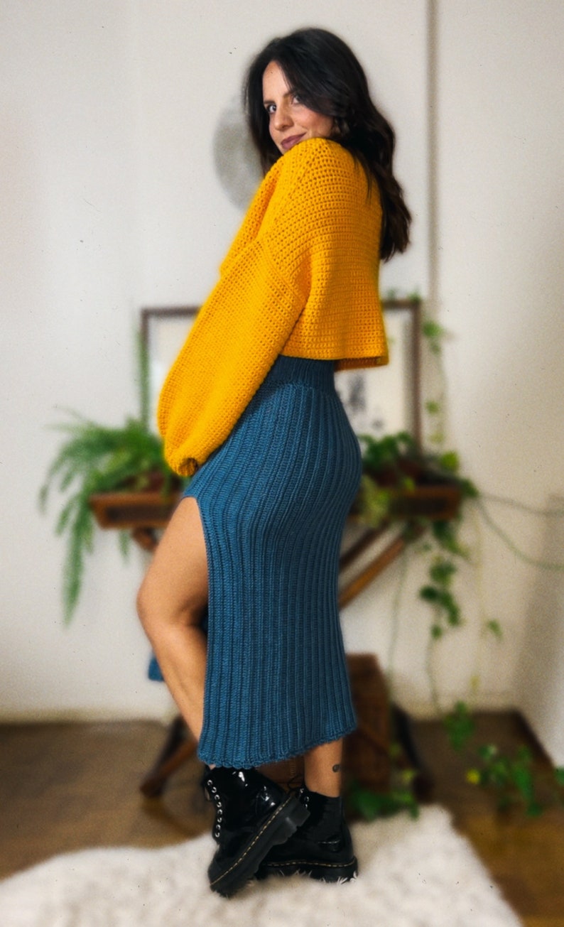 May include: A woman wearing a yellow crocheted sweater and a teal blue crocheted skirt with a slit up the side. She is wearing black leather boots.