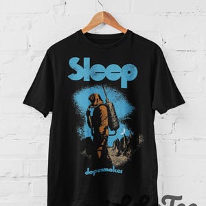 May include: A black short-sleeve t-shirt featuring a graphic print of a hooded figure carrying a large tank on their back. The text "Sleep" appears in large light blue letters above the figure, and the word "dopesmoker" is printed below in blue.