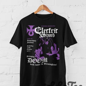 Puede incluir: Una camiseta negra de manga corta con un estampado gráfico en morado y blanco. El diseño incluye el texto Electric Wizard, Capsule Presents, Live at Supersonic Festival, Saturday 22nd October 2011 y Bringing DOOM back home to Birmingham.