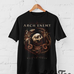 Könnte beinhalten: Ein schwarzes Kurzarm-T-Shirt mit einem Grafikdruck, der den Text ARCH ENEMY und WILL TO POWER zeigt. Das Design zeigt einen zentralen menschlichen Schädel, umgeben von einem Wolfskopf, einem Ziegenkopf und einer Schlange in Braun- und Rottönen.