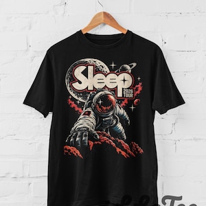 Può includere: Una t-shirt nera con la grafica di un astronauta che si protende su una superficie rocciosa rossa. Sopra l'astronauta, la parola Sleep è stampata in grandi lettere bianche con il testo 2026 TOUR sotto, su uno sfondo di luna e stelle.