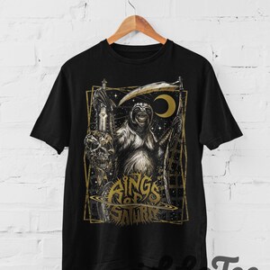 Op de afbeelding: Een zwart t-shirt met korte mouwen en een grafische print van een luiaard verkleed als de dood, met een zeis en een schedel. Het ontwerp bevat een halve maan, sterren en de tekst Rings of Saturn in een goudkleurig, gestileerd lettertype.