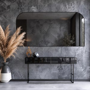 May include: A large arched wall mirror with a thin black frame hangs above a rectangular black console table. A tall vase with dried pampas grass, a gold geometric sculpture, and a black faceted object sit on the console table surface.