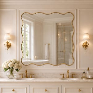 May include: A wall-mounted mirror with a gold frame featuring a wavy, scalloped edge. The mirror is positioned above a double vanity with two gold taps, a white marble countertop, and a glass vase filled with white hydrangea flowers.