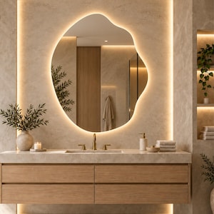 May include: An irregular, organic-shaped wall mirror featuring integrated LED backlighting that emits a warm glow. The mirror is mounted above a light-toned stone countertop with a brass faucet and a wooden vanity cabinet with four drawers.
