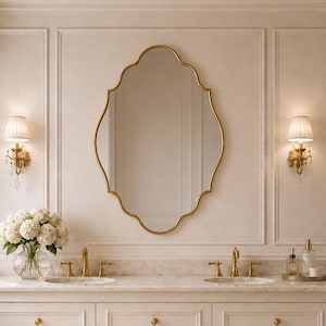 Puede incluir: Un espejo de pared con un marco dorado de bordes festoneados. El espejo se sitúa sobre un tocador doble con dos lavabos, grifos dorados, dos apliques de pared con pantallas blancas plisadas y un florero de cristal con hortensias blancas.