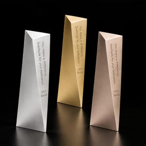 May include: Three triangular awards in silver, gold, and rose gold finishes. Each award features the engraved text: You make a difference, Gratitude for your dedication, 2026, and the name Sarah. The objects have a smooth, metallic surface.