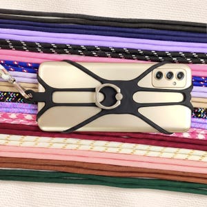 May include: A gold-toned smartphone secured in a black silicone cross-body harness featuring a central circular metal ring stand. The device is positioned over a collection of various colored and patterned fabric lanyards arranged in parallel rows.