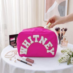 May include: A bright fuchsia makeup bag featuring the name WHITNEY in large, white, varsity-style letters with gold glitter trim. The bag has a top zipper closure and is constructed from a smooth, solid-colored fabric material.