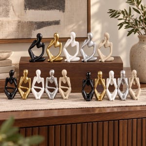 Könnte beinhalten: Eine Sammlung von fünfzehn abstrakten menschlichen Figuren in verschiedenen Posen mit glatten Oberflächen in Schwarz, Gold, Weiß, Silber und strukturiertem Beige. Jede Skulptur zeigt eine stilisierte Person, die mit den Händen am Kopf sitzt.