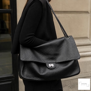 Peut inclure: Un grand sac à bandoulière noir à texture grainée, doté d'un large rabat, d'une fermeture à verrou tournant rectangulaire argentée et d'une fine bandoulière. Le logo blanc NOVIRA BAG est visible dans le coin inférieur droit de l'image.
