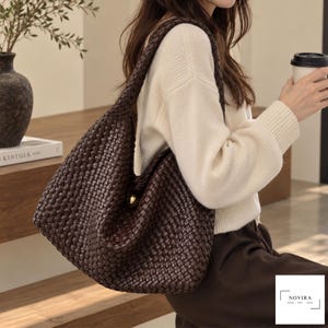 May include: A dark brown hobo bag featuring an intricate woven leather pattern and a single shoulder strap. The bag is secured with a prominent, polished gold-tone circular button closure at the centre. The design is slouchy and unstructured.